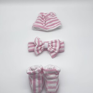 May include: A pink and white striped baby hat, headband, and booties set. The hat is folded in half and the headband has a bow. The booties are made of soft fabric.