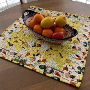 May include: A quilted placemat with a yellow sunflower design and a border featuring farm animals and the word "Farmers". A blue and white bowl filled with oranges, lemons, and a red tomato sits on the placemat, on a wooden table.