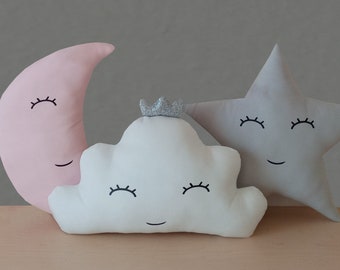 May include: Three plush pillows shaped like a crescent moon, a cloud, and a star. The moon is pink, the cloud is white, and the star is gray. Each pillow has embroidered eyes and a smiling mouth.