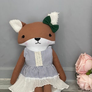 May include: A brown fox plush toy wearing a blue and white striped dress with a white lace trim and a green bow. The fox is sitting on a white platform.
