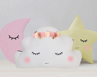 May include: Three plush pillows shaped like a crescent moon, a cloud, and a star. The moon is pink, the cloud is white with a flower crown, and the star is yellow. All three pillows have embroidered eyes and mouths.