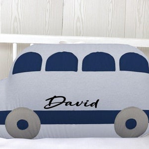 May include: A gray and blue bus-shaped pillow with the name "David" embroidered on it. The pillow is in a crib.