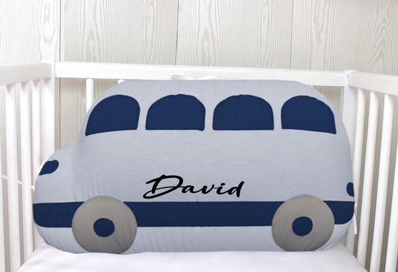 May include: A gray and blue bus-shaped pillow with the name "David" embroidered on it. The pillow is in a crib.