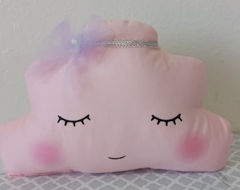 May include: A pink cloud-shaped pillow with a smiling face, closed eyes, and a purple bow with a silver trim.
