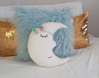 May include: A white crescent moon shaped pillow with a smiling face and eyelashes. The pillow has a blue and pink flower crown on top. The pillow is on a white bed with other pillows.