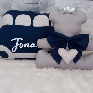 May include: A gray plush teddy bear wearing a silver crown and a blue bow. The bear is sitting next to a blue and gray bus pillow with the name "Jonah" embroidered on it.