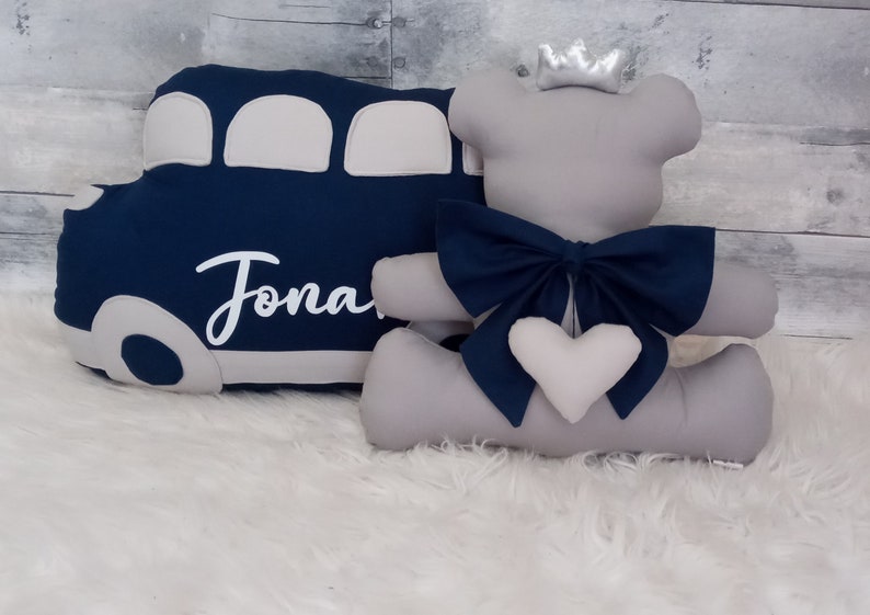 May include: A gray plush teddy bear wearing a silver crown and a blue bow. The bear is sitting next to a blue and gray bus pillow with the name "Jonah" embroidered on it.