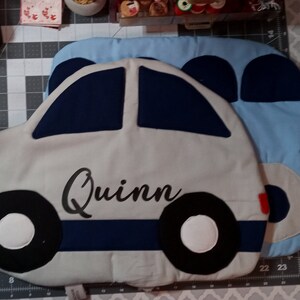 May include: A gray car-shaped pillow with blue and black accents. The pillow has a yellow circle on the front and the name "Quinn" embroidered in black.