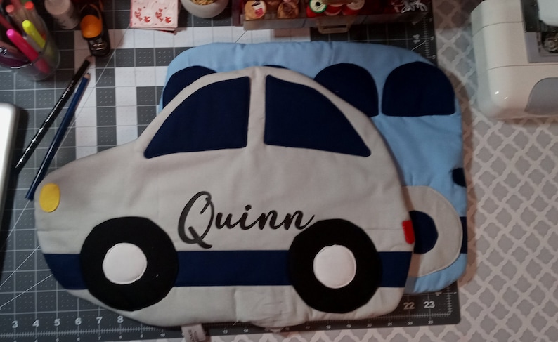 May include: A gray car-shaped pillow with blue and black accents. The pillow has a yellow circle on the front and the name "Quinn" embroidered in black.