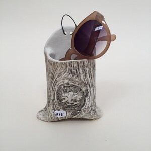 May include: White ceramic tree stump shaped sunglasses holder with a brown and white wood grain pattern. A pair of brown sunglasses are resting in the holder. The number 218 is written on the holder.