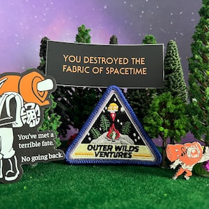 May include: A collection of Outer Wilds merchandise, including a patch with the text "OUTER WILDS VENTURES" and a black sign that reads "YOU DESTROYED THE FABRIC OF SPACETIME". Also includes two character stickers.