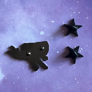 May include: Back view of a black enamel pin featuring a cartoon-style flying creature with a star-shaped pin back. The pin is on a purple background with white speckles.