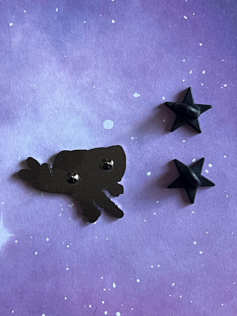May include: Back view of a black enamel pin featuring a cartoon-style flying creature with a star-shaped pin back. The pin is on a purple background with white speckles.