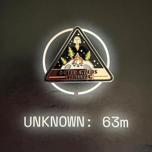 Può includere: Spilla triangolare in smalto con bordo dorato e la scritta "OUTER WILDS VENTURES". La spilla presenta un falò, alberi e un razzo. Lo sfondo è nero con un cerchio bianco. Il testo "UNKNOWN: 63m" è sotto.