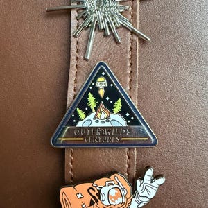 Op de afbeelding: Drie emaille pins op een bruin leren oppervlak. De pins bevatten een zilveren burst design, een driehoekige pin met een kampvuur scène en de tekst "OUTER WILDS VENTURES", en een duiker karakter in oranje en wit.