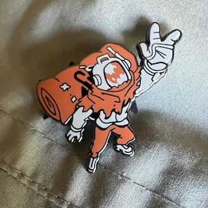May include: Orange and white enamel pin of a cartoon astronaut wearing a spacesuit with a rolled-up sleeping bag on their back. The astronaut is waving with their right hand.