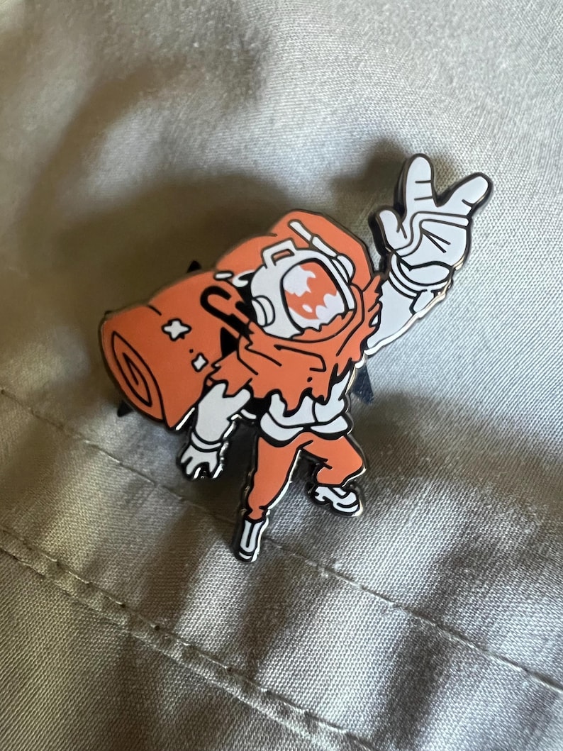 May include: Orange and white enamel pin of a cartoon astronaut wearing a spacesuit with a rolled-up sleeping bag on their back. The astronaut is waving with their right hand.