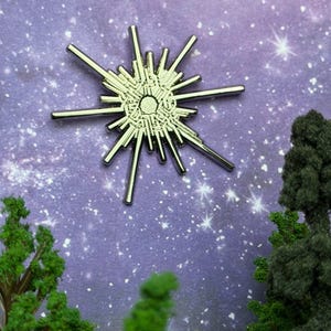 May include: A silver pin badge in the shape of a starburst or sun, featuring radiating lines and a detailed central design. The pin is set against a purple background with a starry night sky and miniature trees.
