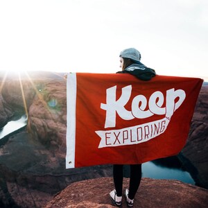 以下が含まれることがあります： 白い文字で「Keep Exploring」と書かれた赤い旗を持った人がいます。旗は風になびいています。その人は、峡谷を見下ろす岩の崖の上に立っています。