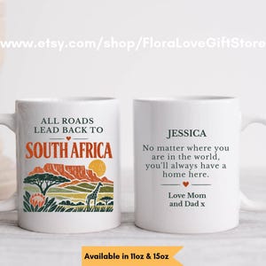 Peut inclure: Deux tasses en céramique blanche avec des motifs imprimés. La tasse de gauche montre un paysage avec une girafe, un arbre et le texte "ALL ROADS LEAD BACK TO SOUTH AFRICA". La tasse de droite porte le nom "JESSICA" et un message personnalisé.