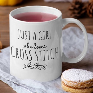 May include: White ceramic mug with a black design that reads "Just a girl who loves cross stitch". The mug is filled with a pink beverage and has a white handle. The mug is sitting on a gray linen napkin with a small, round, powdered cookie next to it.