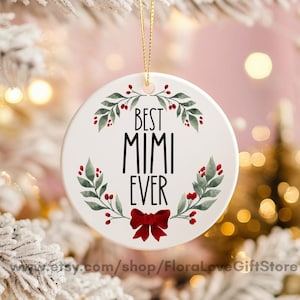 Puede incluir: Adorno navideño de cerámica blanca con el texto negro "BEST MIMI EVER". El adorno está decorado con hojas verdes, bayas rojas y un lazo rojo. Se adjunta una cuerda dorada para colgarlo.