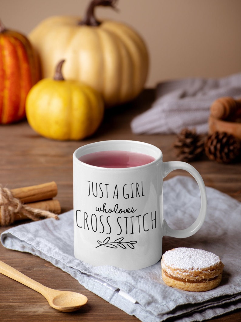 May include: White ceramic mug with a black design that reads "Just a girl who loves cross stitch". The mug is filled with a pink beverage and has a white handle. The mug is sitting on a gray linen napkin with a small, round, powdered cookie next to it.