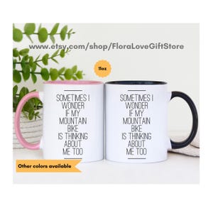 May include: Two 11-ounce white ceramic mugs with black handles. The mug on the left has a pink rim and the mug on the right has a black rim. Both mugs have the same text printed in black: "Sometimes I wonder if my mountain bike is thinking about me too."