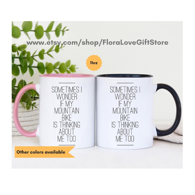 May include: Two 11-ounce white ceramic mugs with black handles. The mug on the left has a pink rim and the mug on the right has a black rim. Both mugs have the same text printed in black: "Sometimes I wonder if my mountain bike is thinking about me too."