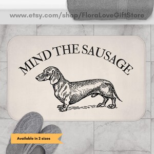 May include: Cream-colored bath mat with the text "MIND THE SAUSAGE" above a black and white illustration of a dachshund. The mat is rectangular with rounded corners. The text "Available in 2 sizes" is also present.