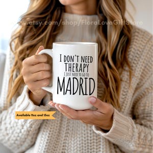 Op de afbeelding: Een witte keramische mok met de zwarte tekst I DON'T NEED THERAPY I JUST NEED TO GO TO MADRID. De mok heeft een handvat aan de zijkant en is beschikbaar in 11oz en 15oz formaten, zoals aangegeven op het oranje label linksonder.