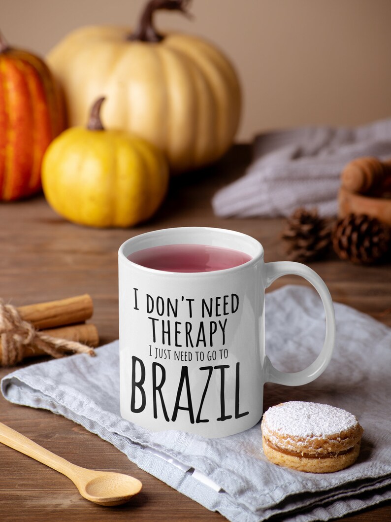 Pu&ograve; includere: Una tazza in ceramica bianca con una grafica nera che recita "Non ho bisogno di terapia, ho solo bisogno di andare in Brasile". La tazza &egrave; piena di una bevanda rosa e si trova su un tovagliolo di lino grigio con un cucchiaio di legno e un biscotto.