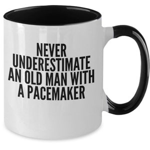 May include: White ceramic mug with a black interior, handle, and rim. The mug features the text "NEVER UNDERESTIMATE AN OLD MAN WITH A PACEMAKER" in bold black letters. A humorous gift idea.