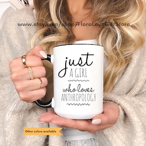 Può includere: Una tazza in ceramica bianca con manico e bordo neri, caratterizzata dalla scritta stampata in nero 'just A GIRL who loves ANTHROPOLOGY' con linee ondulate decorative sopra e sotto la parola 'who loves'. La tazza è tenuta da due mani.