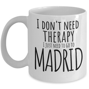 K&ouml;nnte beinhalten: Wei&szlig;e Keramik-Kaffeetasse mit schwarzem Text, der "Ich brauche keine Therapie, ich muss nur nach Madrid" lautet.