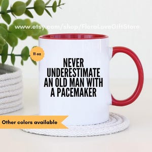 May include: White ceramic mug with a red rim and handle. The mug features a black graphic that reads "Never underestimate an old man with a pacemaker". The mug is 11 oz and is sitting on a white rope coaster.
