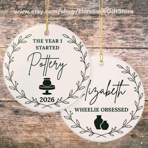 May include: Two round white ceramic ornaments with gold hanging strings. The first features a black leaf wreath, the text THE YEAR I STARTED Pottery 2026, and a pottery wheel icon. The second features a leaf wreath, the name Elizabeth, the text WHEELIE OBSESSED, and two pottery vase icons.