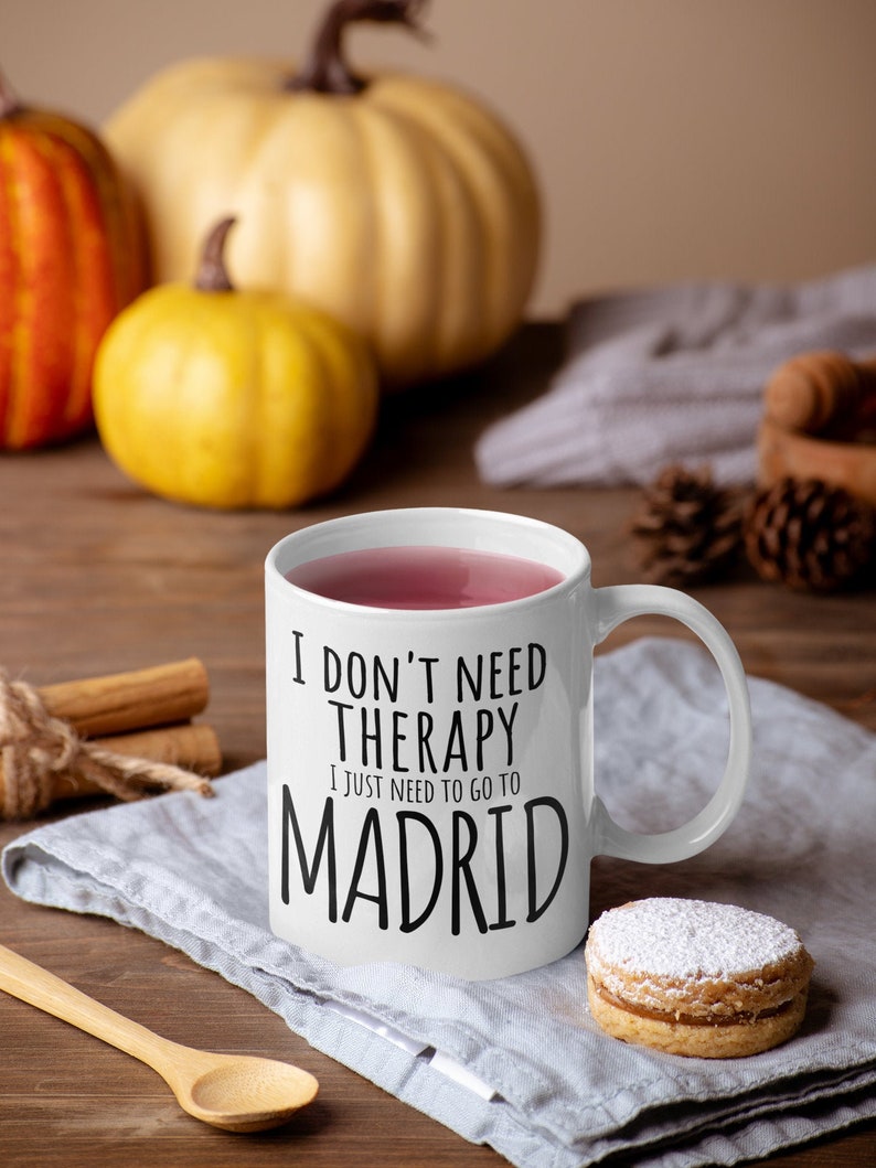K&ouml;nnte beinhalten: Eine wei&szlig;e Keramiktasse mit schwarzem Text, der "Ich brauche keine Therapie, ich muss nur nach Madrid" lautet. Die Tasse ist mit einem rosa Getr&auml;nk gef&uuml;llt und steht auf einer grauen Leinen-Serviette mit einem Keks daneben.