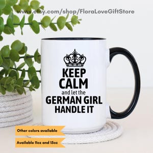 May include: White ceramic mug with a black rim and handle. The mug has a black graphic that says "Keep Calm and let the German Girl Handle It" with a crown above the text. The mug is sitting on a white rope coaster.