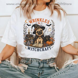Könnte beinhalten: Ein weißes T-Shirt mit einer Grafik eines Mopses, der einen Hexenhut und einen Umhang trägt, umgeben von Büchern, einer Kerze, einem Zaubertrank, einem Kürbis und einem Geist. Der Text lautet WRINKLES & WITCHCRAFT in orangefarbenen und schwarzen Buchstaben.