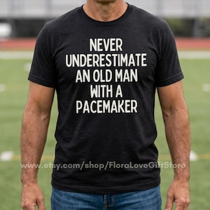 Op de afbeelding: Een antracietgrijs T-shirt met korte mouwen en de witte tekst NEVER UNDERESTIMATE AN OLD MAN WITH A PACEMAKER op de voorkant. Het shirt heeft een ronde hals en een gemêleerde stofstructuur en wordt gedragen met een blauwe spijkerbroek.