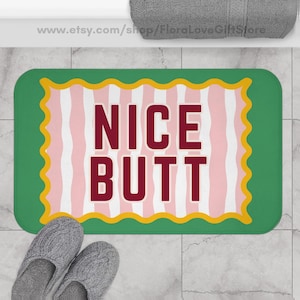 May include: A rectangular bath mat with the words "NICE BUTT" in burgundy block letters. The mat has a green border with a yellow scalloped edge. The center features pink and white vertical stripes.