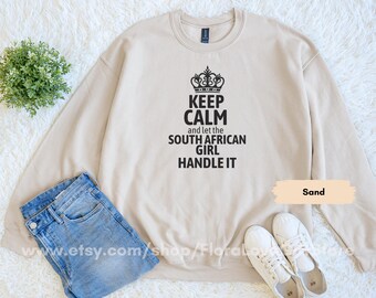 Op de afbeelding: Een zandkleurige sweatshirt met de zwarte tekst "KEEP CALM and let the SOUTH AFRICAN GIRL HANDLE IT". De afbeelding toont ook een blauwe jeans en witte sneakers, alles op een witte, getextureerde achtergrond.