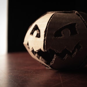 K&ouml;nnte beinhalten: Ein K&uuml;rbis aus Pappe mit einem geschnitzten Jack-o'-Lantern-Gesicht. Der K&uuml;rbis ist braun und hat einen dunklen Hintergrund.