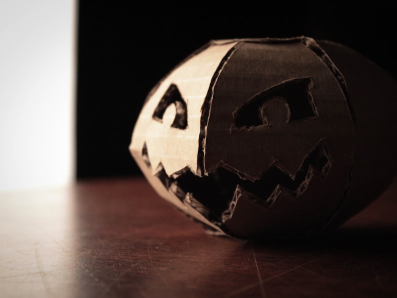 K&ouml;nnte beinhalten: Ein K&uuml;rbis aus Pappe mit einem geschnitzten Jack-o'-Lantern-Gesicht. Der K&uuml;rbis ist braun und hat einen dunklen Hintergrund.