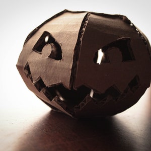 K&ouml;nnte beinhalten: Eine braune Papp-Halloween-Laterne mit einem geschnitzten Gesicht. Die Augen und der Mund sind aus dem Karton ausgeschnitten.