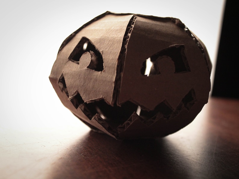 K&ouml;nnte beinhalten: Eine braune Papp-Halloween-Laterne mit einem geschnitzten Gesicht. Die Augen und der Mund sind aus dem Karton ausgeschnitten.