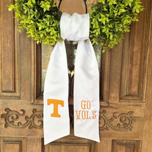 May include: White graduation sash with orange embroidered letters and the words "GO VOLS" and a "T" logo. The sash is hanging on a wooden door with a green wreath.