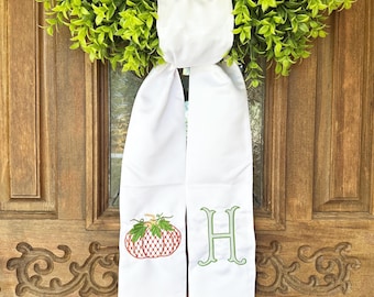 May include: White door sash with embroidered fall-themed designs. Features a red and green pumpkin and a green letter "H". The sash is hanging on a wooden door with a green wreath.