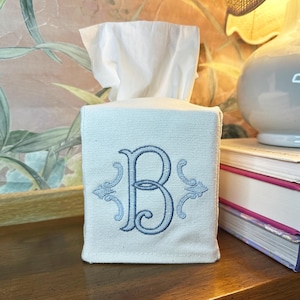 May include: A square, off-white tissue box cover with a light blue embroidered letter "B" and decorative flourishes. White tissues are visible at the top. The cover is made of a textured fabric.